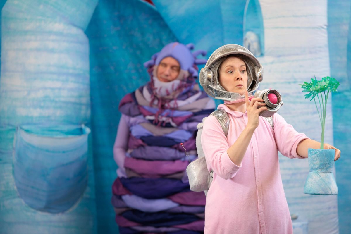 Jem as Baby in Space Neighbours on Uranus, aiming her toy camera at a green plant in a blue pot. Behind her, the purple alien dozes with eyes closed among the icy blue set.