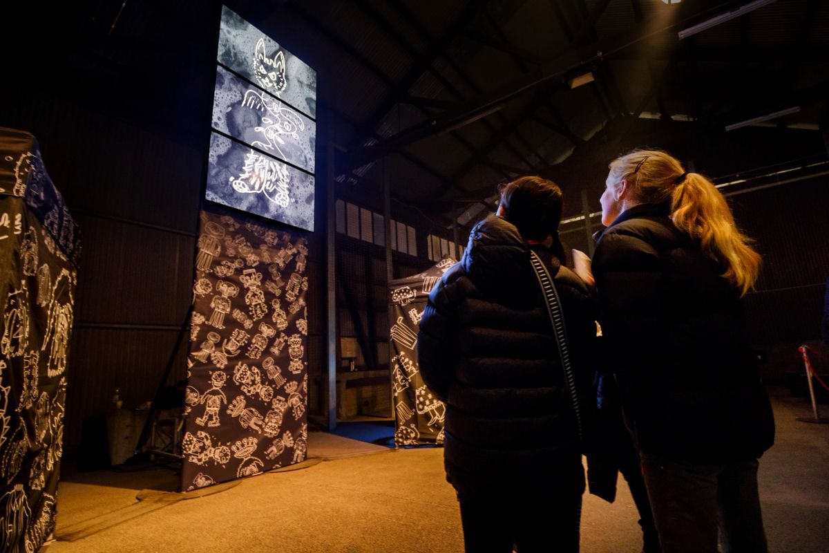 A group of people are standing and looking at black marquees covered in white drawings as well as up high there are three tv screens showing a black background and white drawing