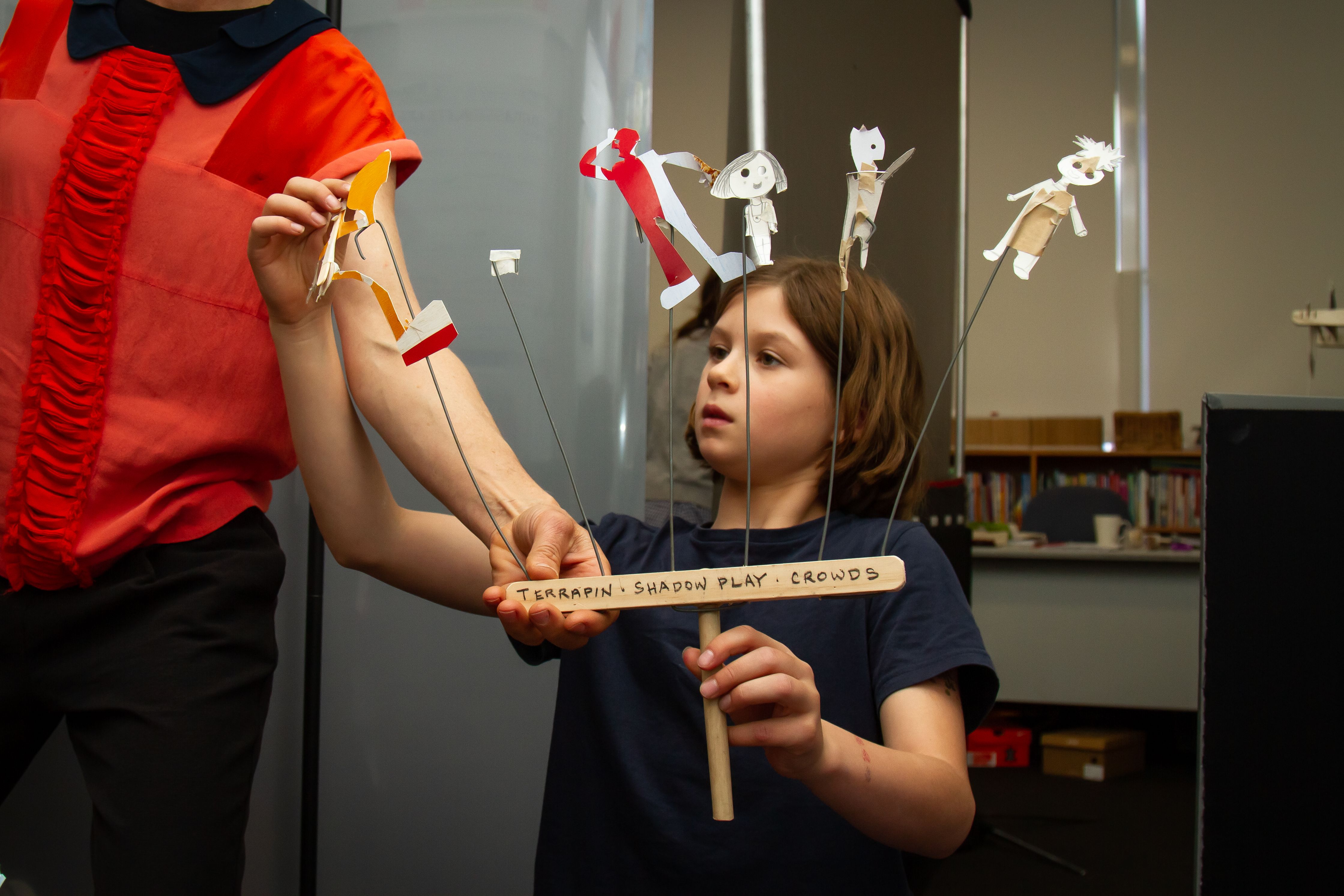 A young person holds up a wooden handle, attached to the handle are six seperate shadow people made from what looks to be cardboard boxes. these people will create shadows and stories with a projector