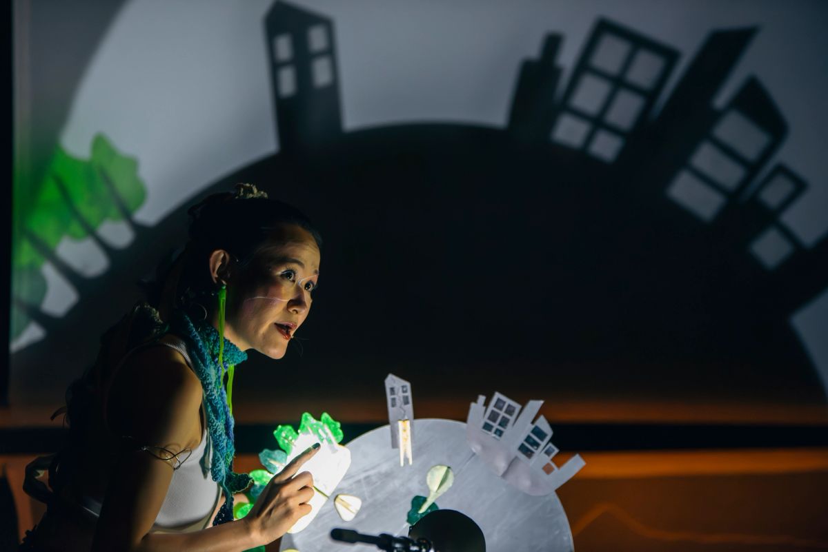 A performer with expressive makeup and a blue scarf gestures while manipulating a small-scale model of a city with paper buildings and green elements. Behind them, a shadow projection of a larger cityscape is cast onto a screen.