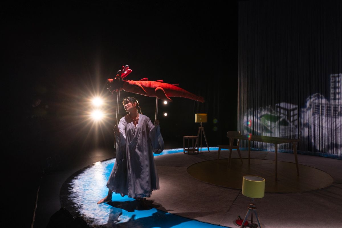 A performer dressed in a flowing robe holds a red dragon puppet under stage lights. She represents the mother in The Story of Chi, performed at Adelaide Festival Centre as part of OzAsia Festival 2024.