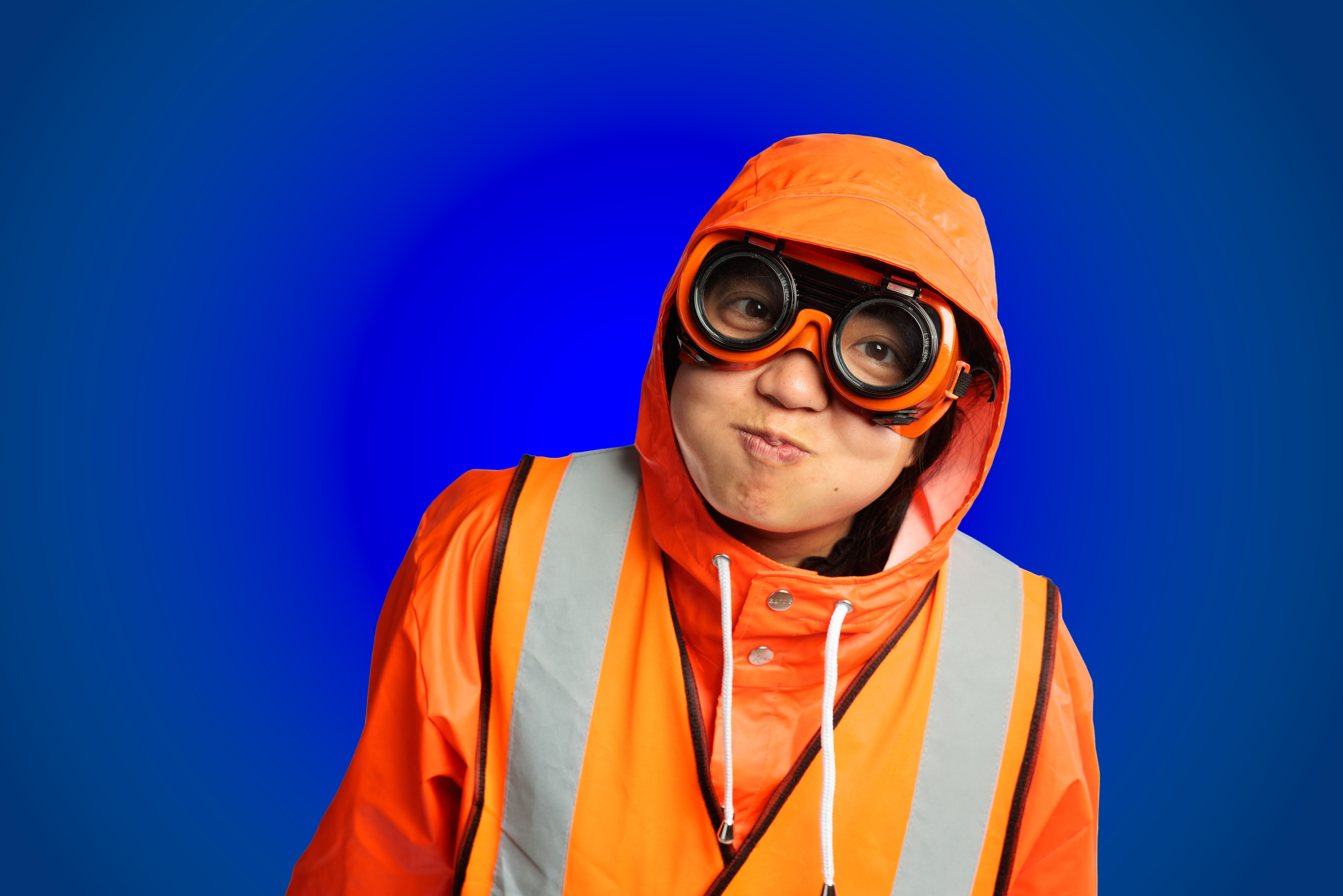 A person wearing an array of fluro orange clothing, goggles and a raincoat is set in front of a bright blue background.
