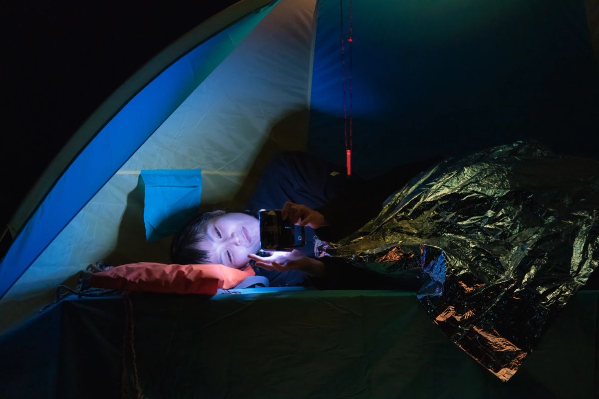 A person lays in a tent, covered by reflective bedding and is on their phone. the only light source is the phone