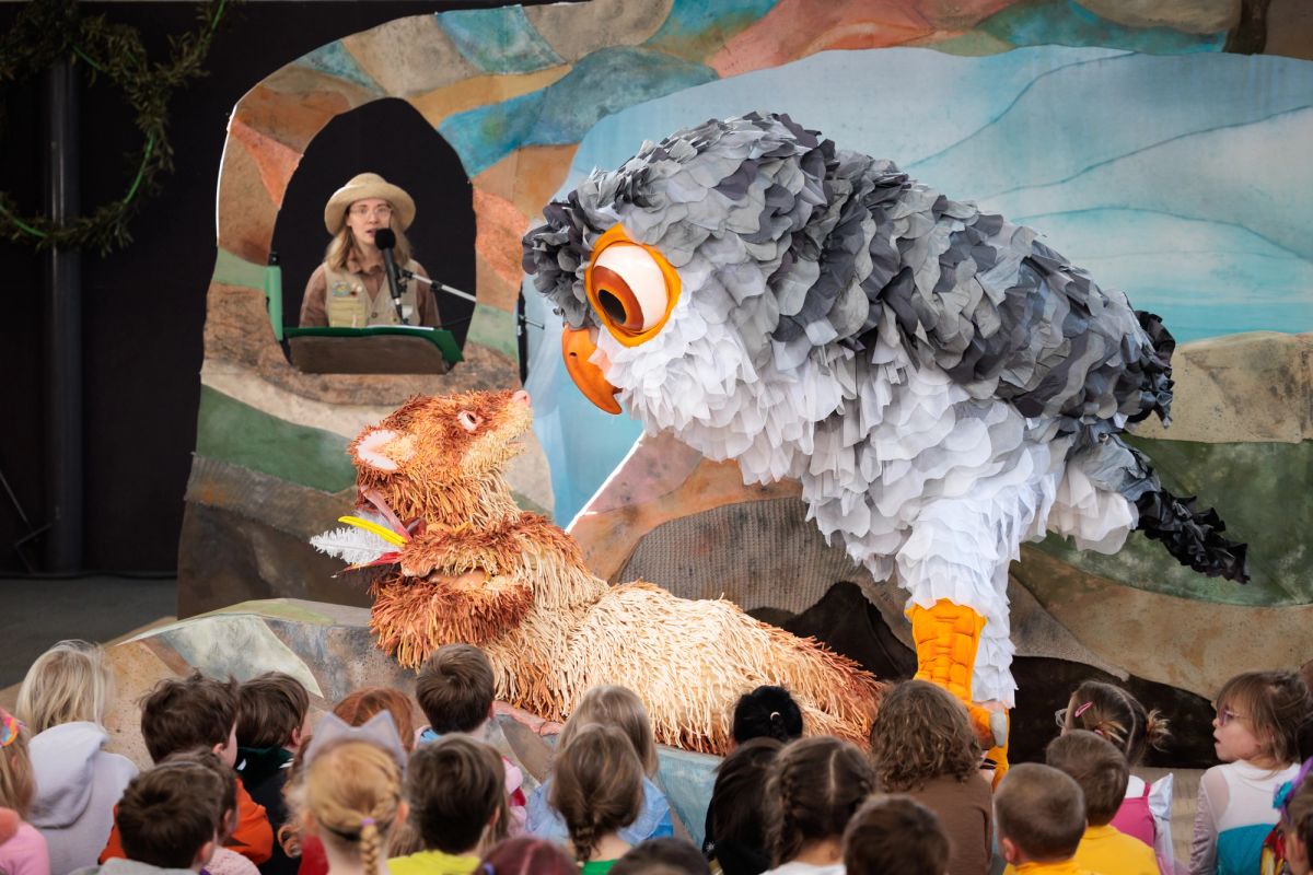 Feathers, a giant puppet ferret lays is in front of a group of school children, a giant owl puppet over the top of the ferret, the set behind Feathers and the owl is nature coloured, green, blue and sandy colour.