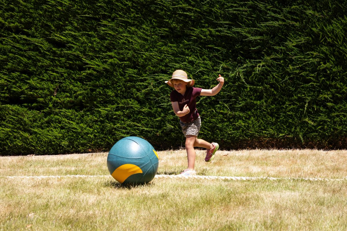 This is a bright sunny day, we see one young person wearing a t-shirt and shorts, as well as a sunhat, playing a game of giant marbles, they’ve just thrown their giant marble and behind them is what looks to be a big green hedge.