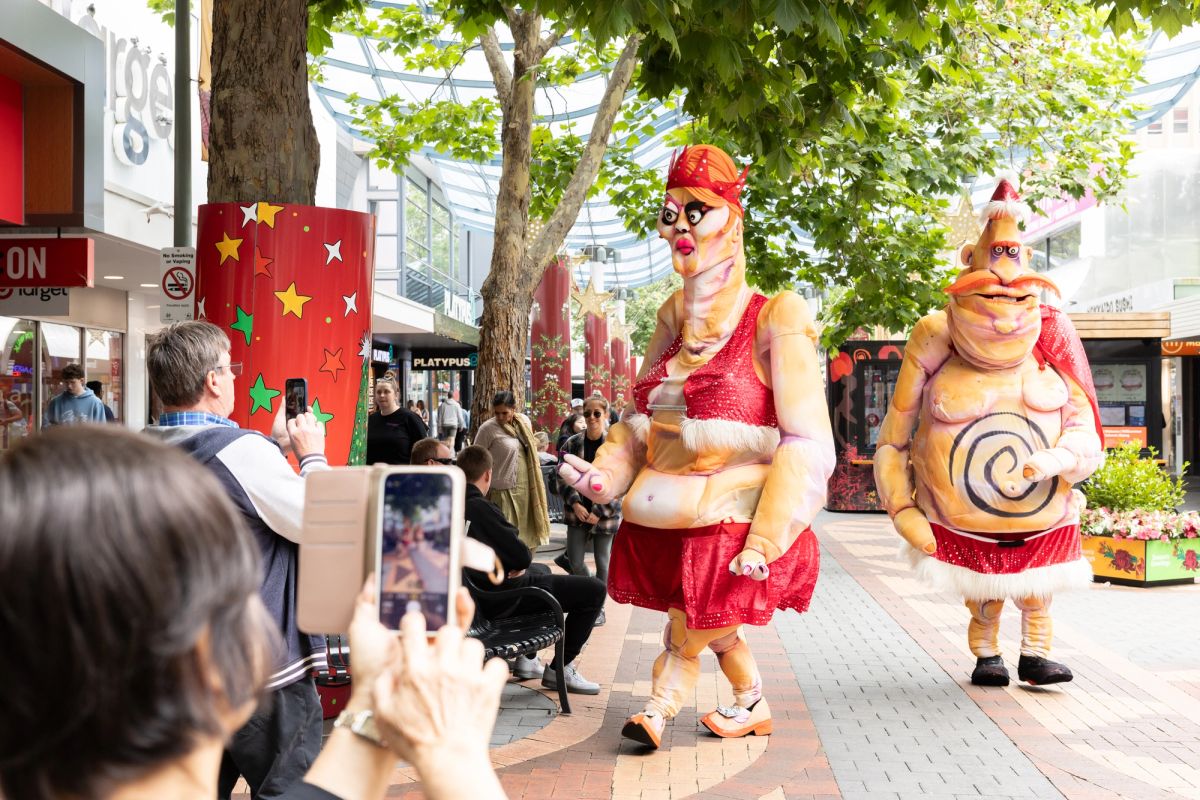 Two giant roving puppets, named Ma and Pa Ubu who both have exaggerated features, they wear Christmas themed outfits, one has a large orange moustache, the other bright neon pink lips, are walking in the City of Hobart Christmas Pageant, 2024.