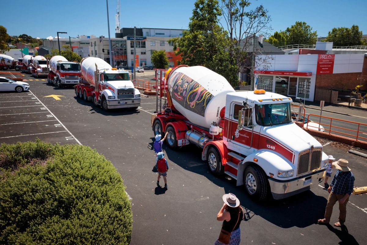 parked on a road on a sunny day is a series of cement mixers, one is very close to the camera and on the side of it is a graphic designed face