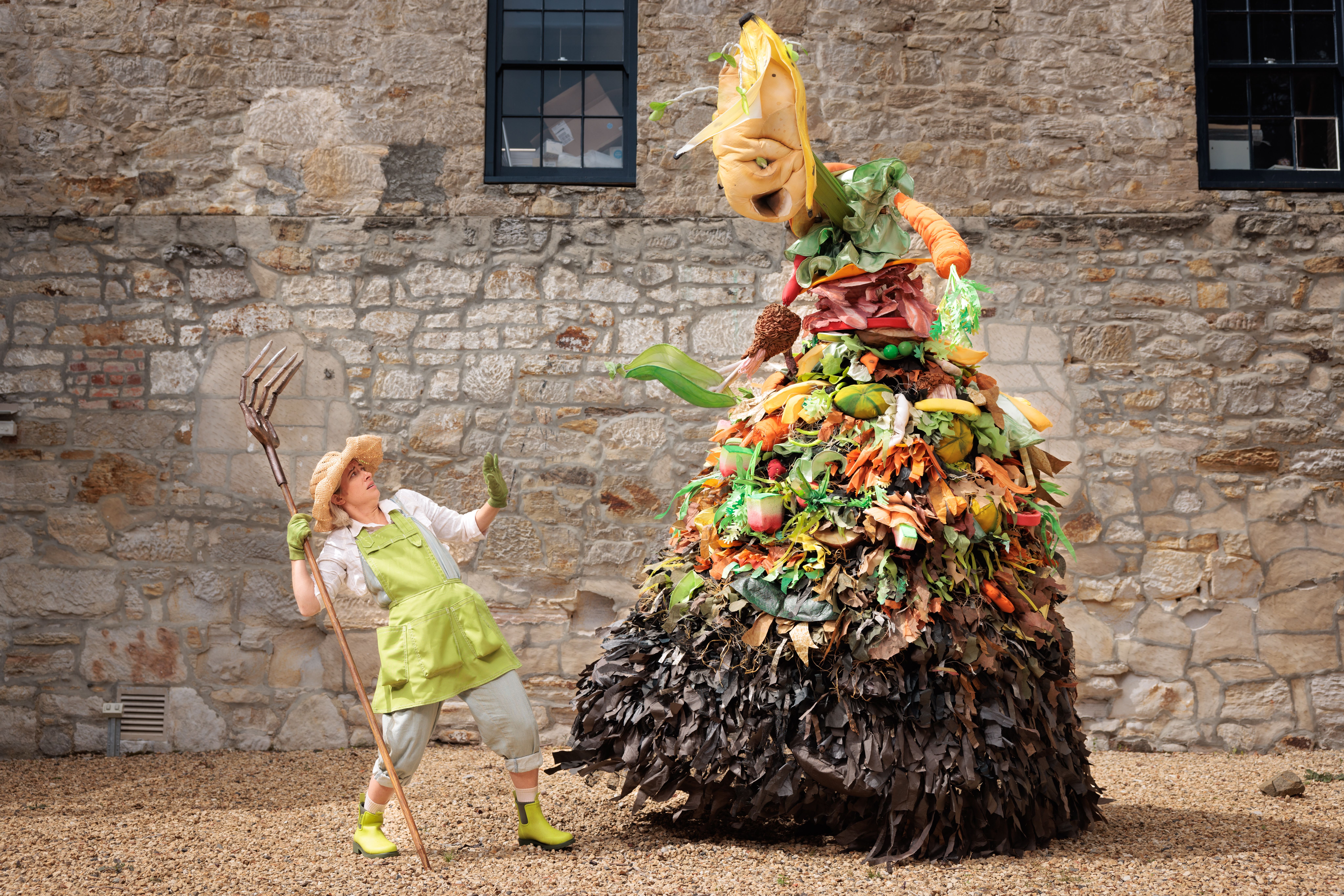 A puppet compost heap made of fabric and foam sits on a yellowy stone ground in front of a sandstone backdrop. There is a person, a gardener holding a pitchfork next to the larger than life compost heap, this gardener looks scared of the puppet.