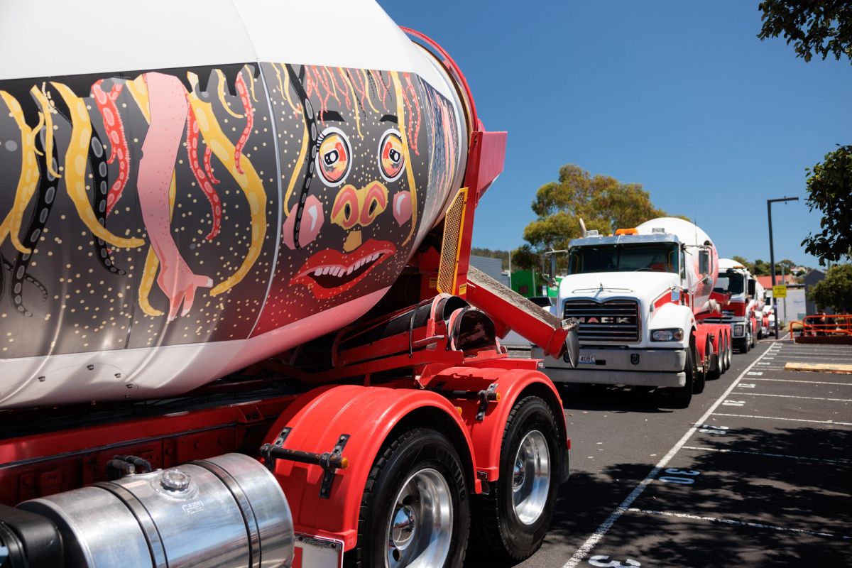 parked on a road on a sunny day is a series of cement mixers, one is very close to the camera and on the side of it is a graphic designed face