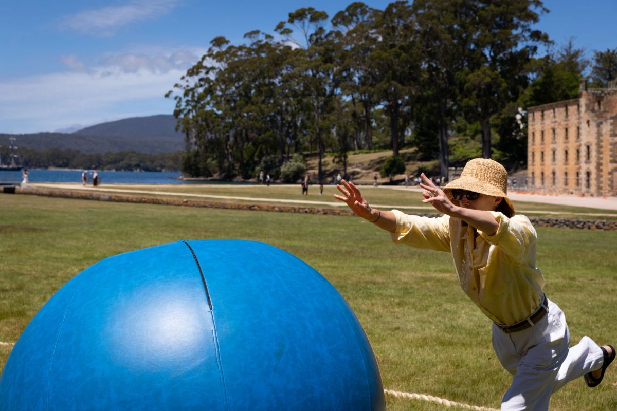 This is a bright sunny day, we see one person, who looks as though they’ve just pushed their giant marble, they are playing a game of giant marbles on the lawn in front of the historic sandstone building of Port Arthur. n the distant background we see people walking and a landscape of a waterway and many big trees.