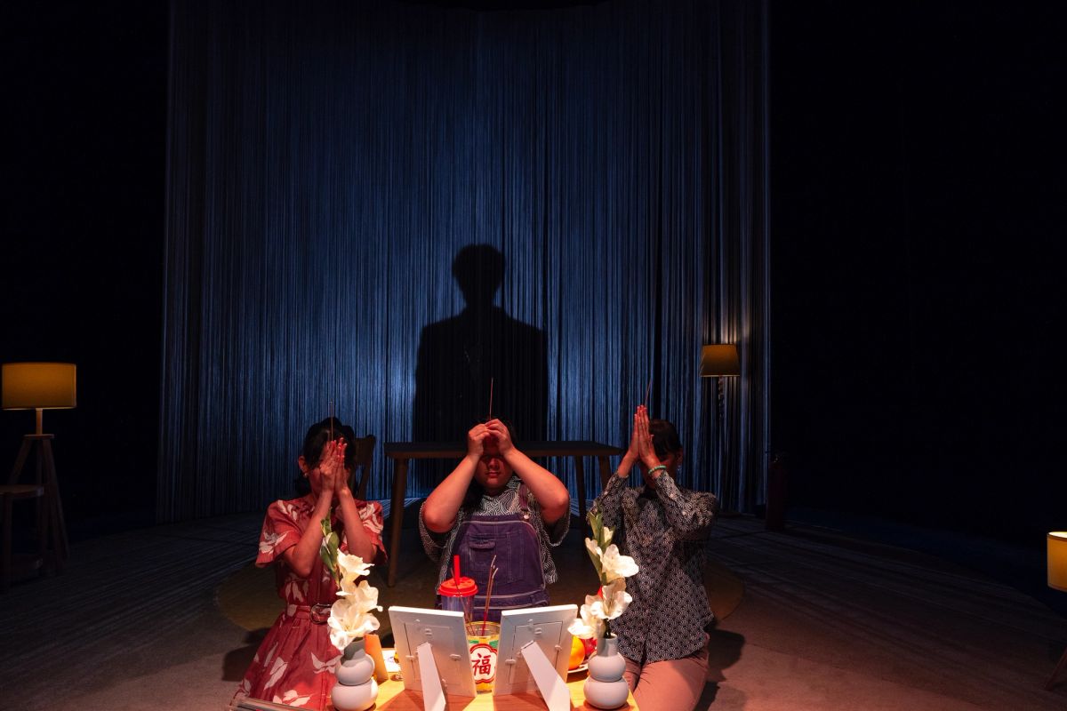 Three performers portraying the grandmother, mother, and daughter kneel at an altar holding incense sticks. Behind them, a shadow of the father is cast on the curtain, in a scene from The Story of Chi, performed at Adelaide Festival Centre as part of OzAsia Festival 2024.