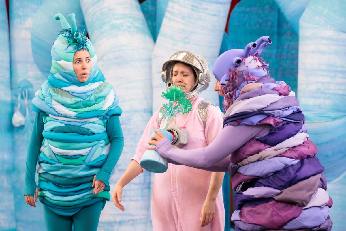 The purple alien holds a potted plant up to Jem as Baby, who screws up her face at the smell, in Space Neighbours on Uranus. The teal alien looks on, wide-eyed.