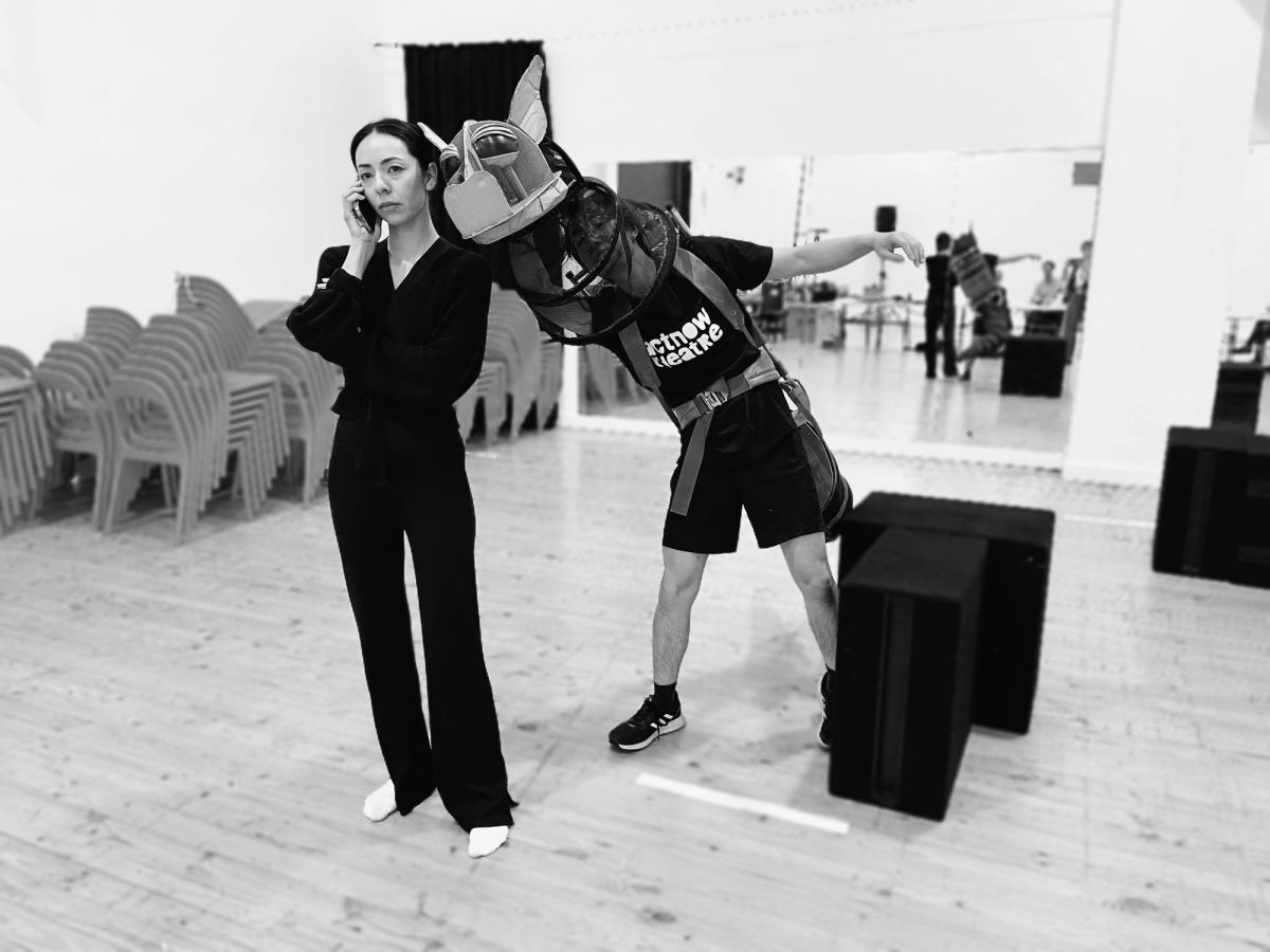 two people in a rehearsal room with wooden floors and chairs stacked in the background stand, one wearing a dragon costume made from cardboard, the other holding their phone to their ear