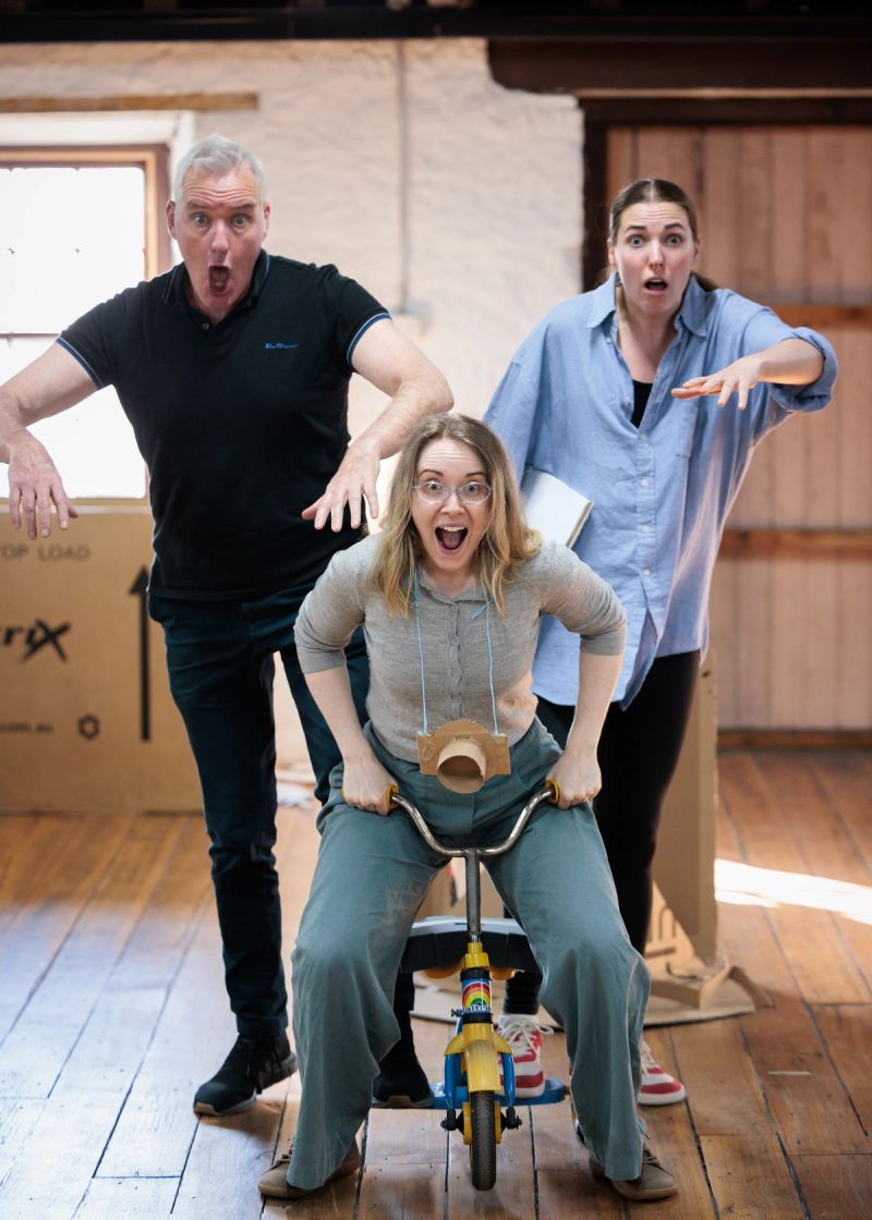 Three people pose playfully in a rehearsal room. One person sits on a small tricycle, gripping the handlebars and leaning forward, while two others stand behind them with exaggerated surprised expressions and raised arms. Cardboard props and a wooden floor are visible in the background.