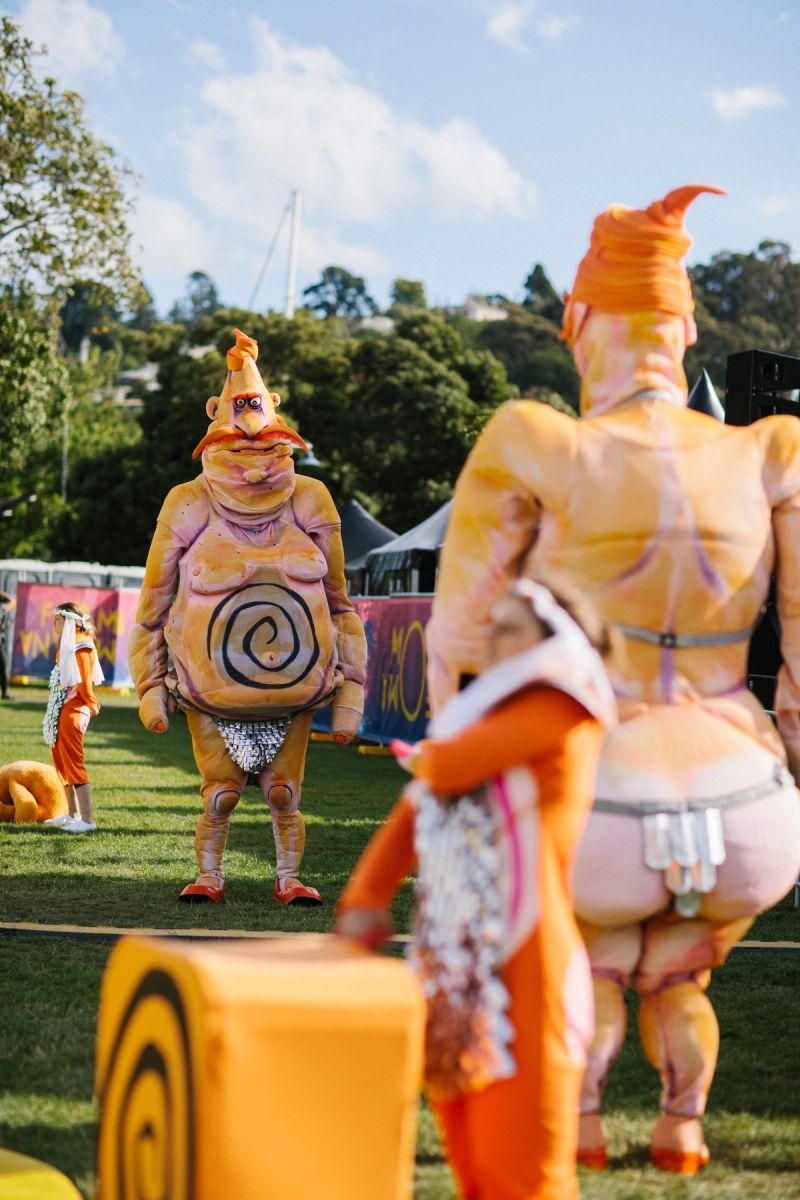 Two larger than life characters, Pa Ubu and Ma Ubu stand looking at one another, we see the back of Ma Ubu and the front of Pa Ubu. They are at a festival site on a bright sunny day, without anyone around them.