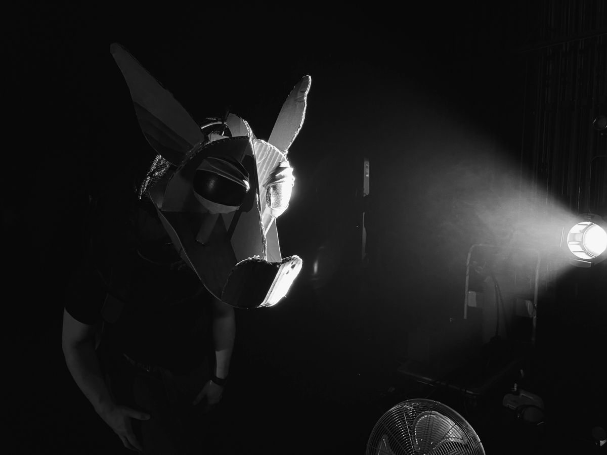 A black and white image of a cardboard dragon head on a person, a light is right beside them lighting one side of the face