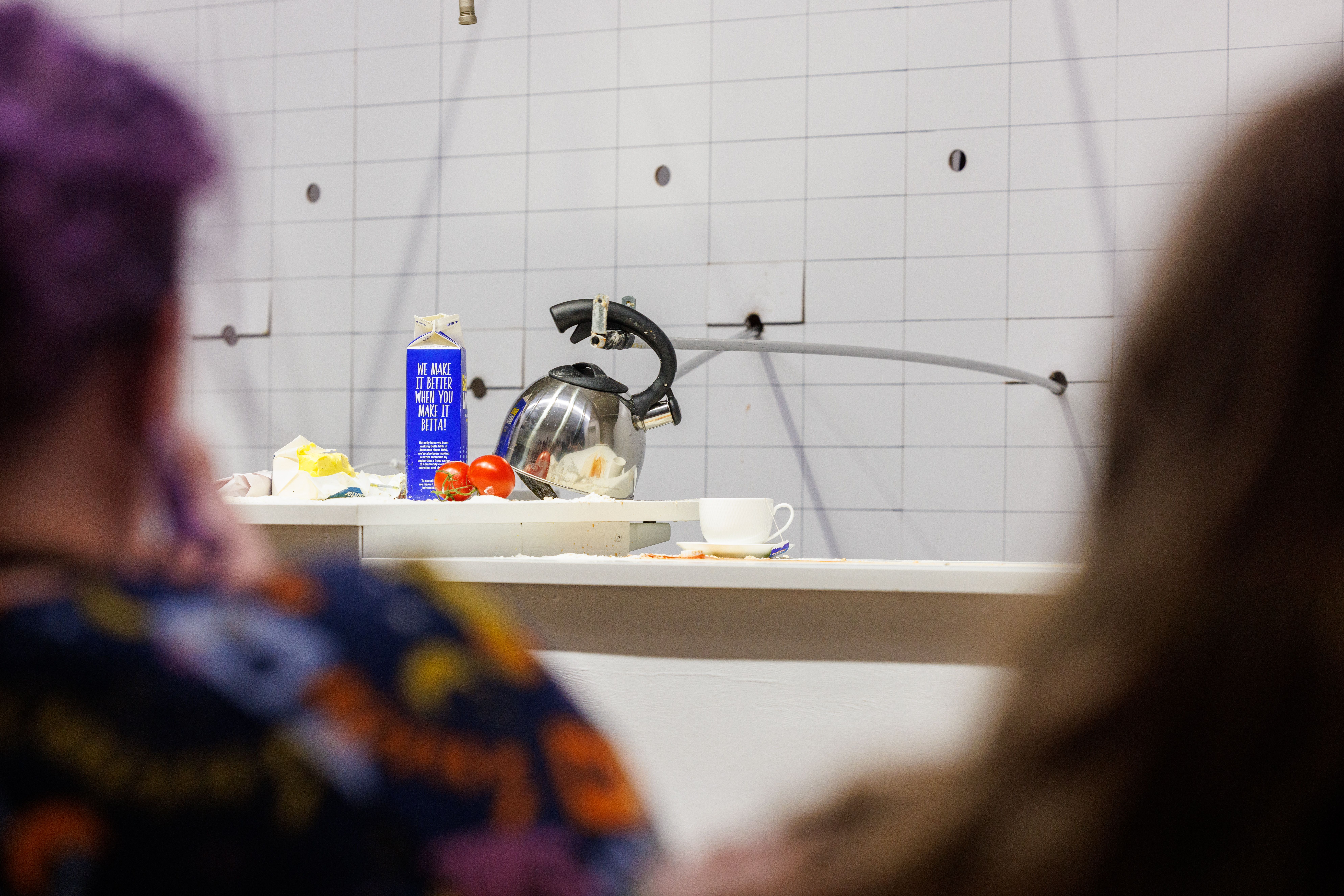 Looking between two people we see a wall, with sticks/poles poking out from the wall, these poles are holding are trying to be the hands for a kettle pouring a cup of tea. We also see two tomatoes, a cup and saucer and a 1litre carton of milk.