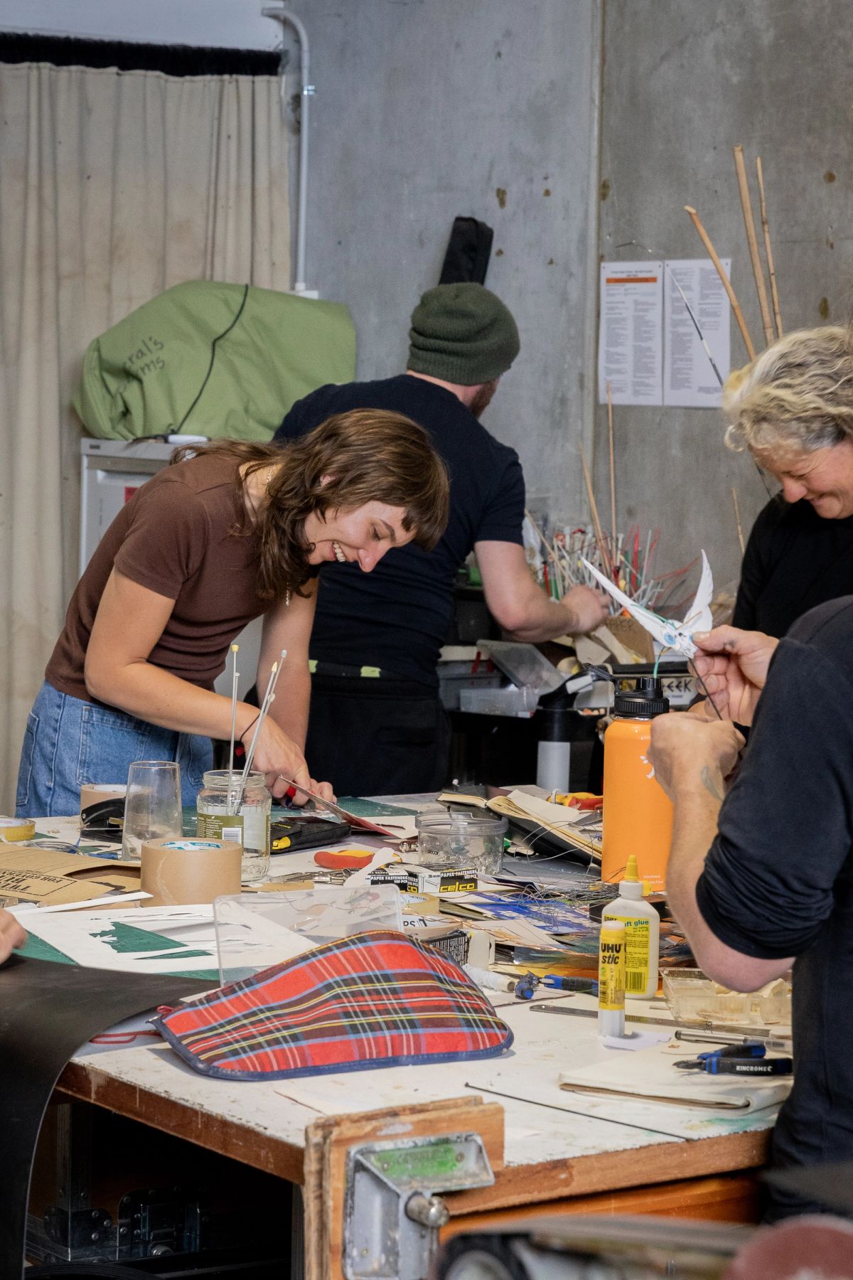Around a work bench are a number of makers, the bench is covered in glue, cardboard, knitting needles and various other things.
