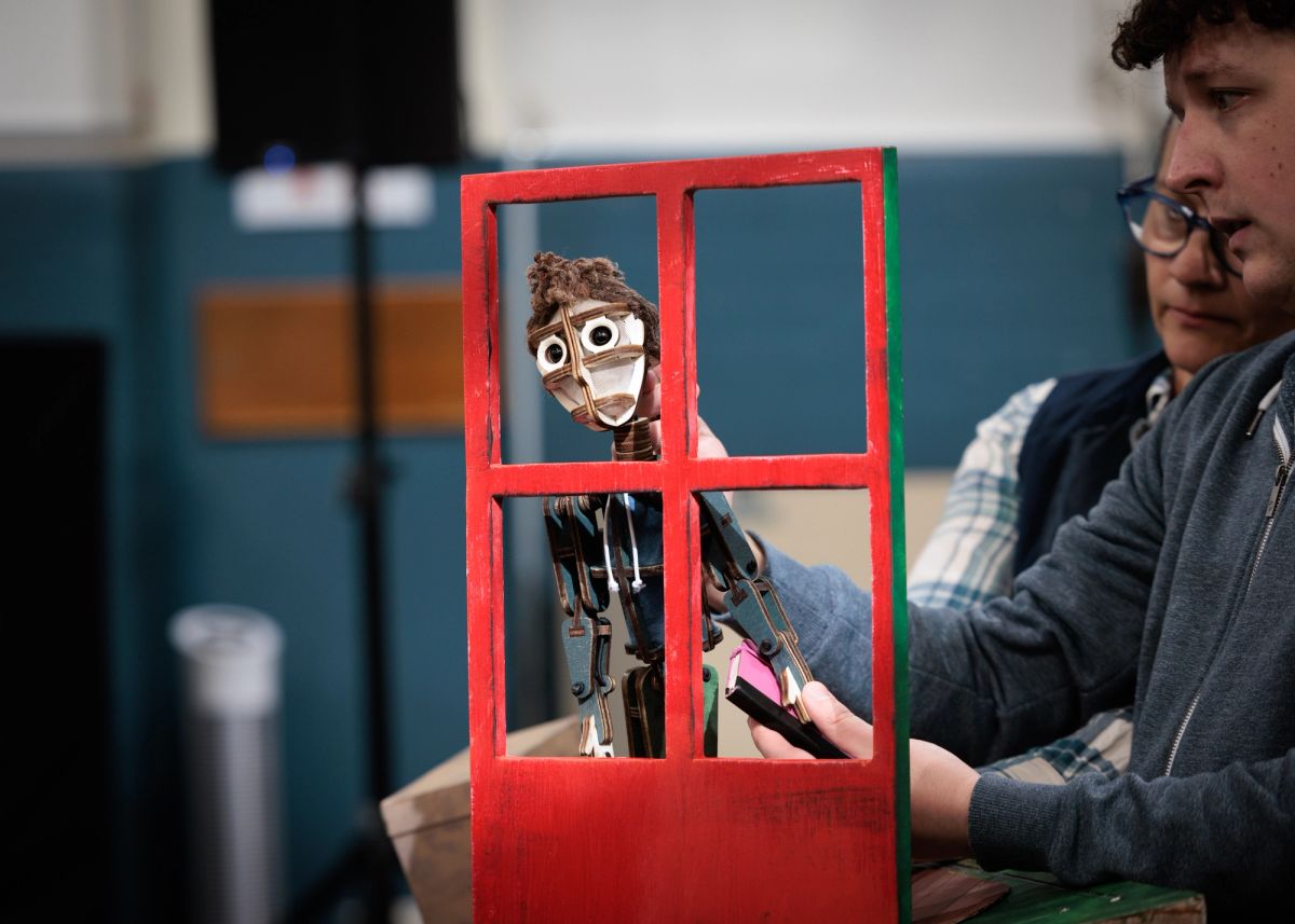 There is a vibrant red door that has four panes where glass could be. A boy puppet is poking his head through the space, looking up to the middle left. A performer holds him there and is in the background.