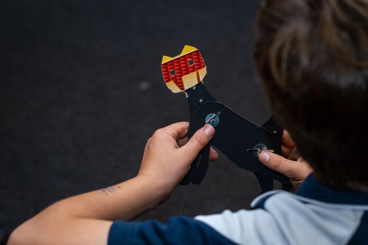 A small child holds a cat like character, we look down over the childs shoulder and see the cat is made from random card board boxes, the head has a pattern of red and yellow whilst the body is all black.