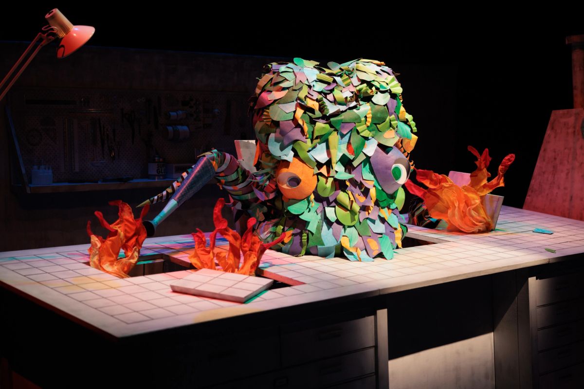 On a white checkerboard table is a giant multicoloured octopus with two eyes and tentacles going off in all the directions, on either side of the octopus is three fires make from fabric poking through the playboard. This scene is under stage lighting.