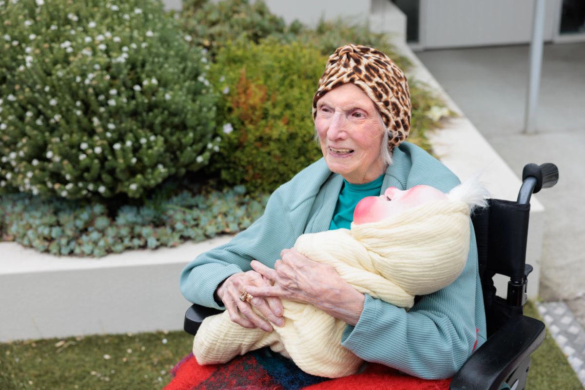 An elderly person sits in a wheelchair, they wear a leopard print head wrap and a blue jumper, they sit and hold a baby puppet