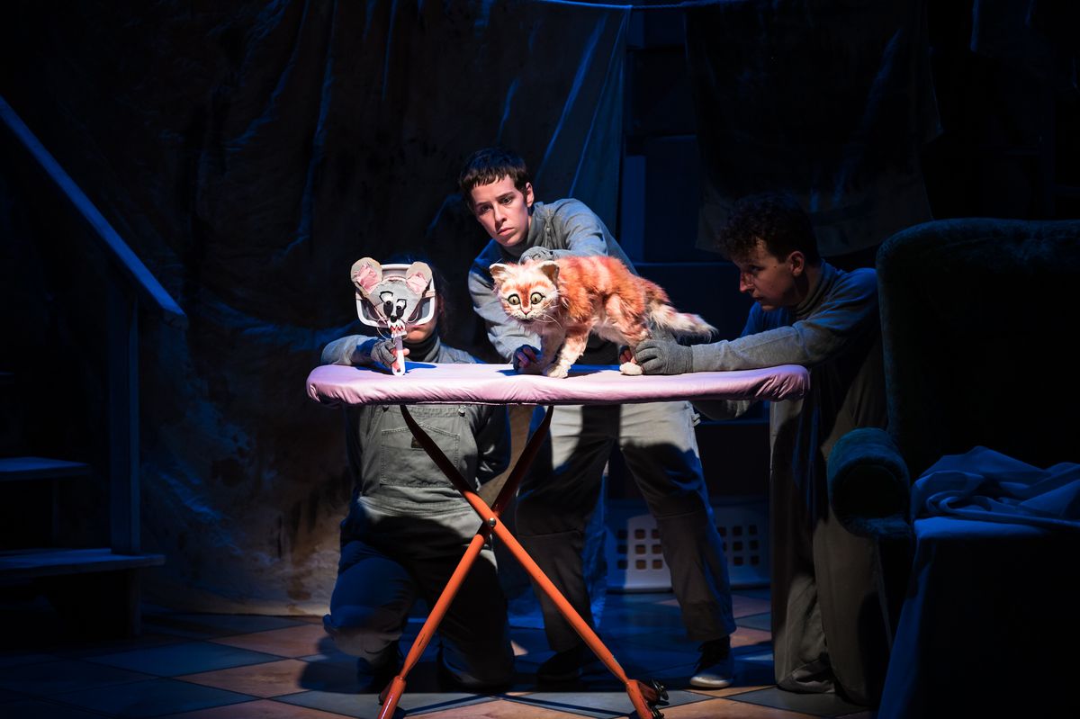 Under stage lights we see an ironing board with orange legs and a pink top, surrounding the ironing table we see three puppeteers in grey boiler suits, holding an orange tabby cat puppet and a mirror which has the face of a mouse taped to it. the surrounding background is dark blue and grey and we're hardly able to make out anything, except a chair and what looks to be stairs