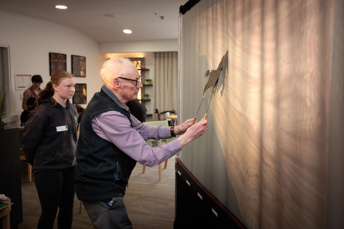 an elderly person holds a shadow puppet horse up to a screen, a light comes from behind to light up the screen. A young person with a name tag stands off the the side of the elderly person.