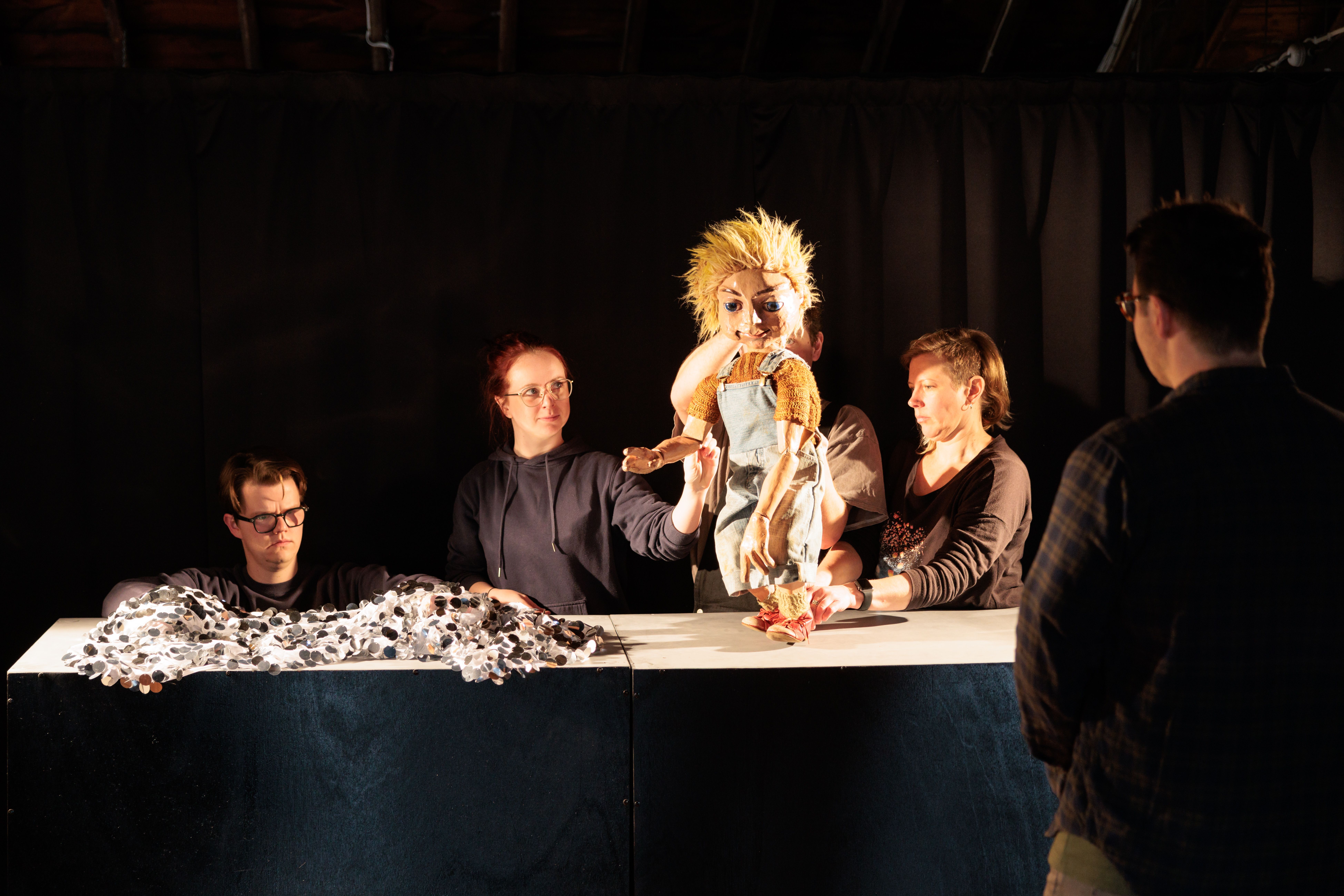A group of people under theatre lights stand at a table on the table there are puppets and off to the side somebody is watching them use the puppets