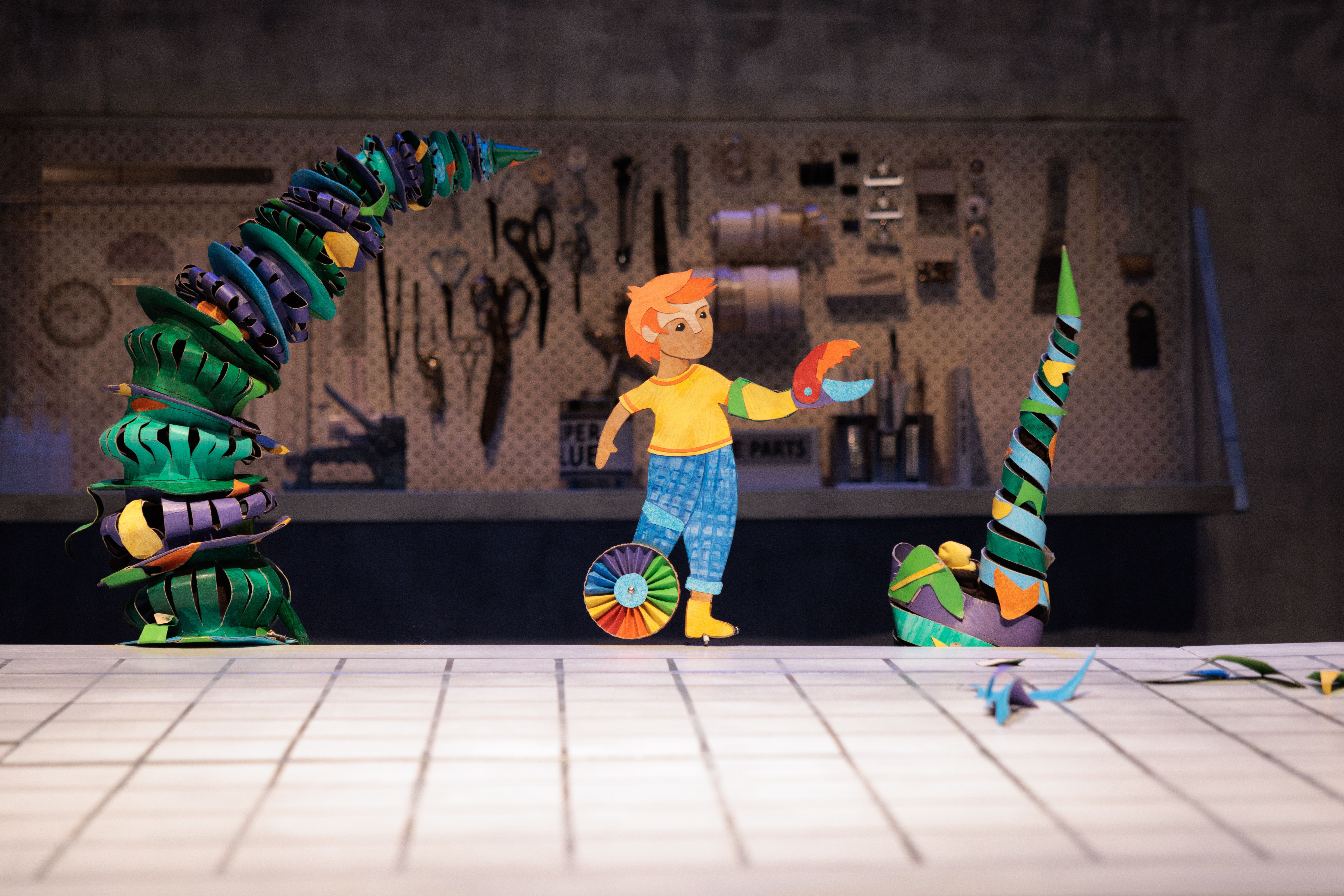 A paper cut out boy called Benny, who has one leg, the other a wheel stands on top of a table, behind him looks to be a workshop and on either side of him are coloured tentacles.