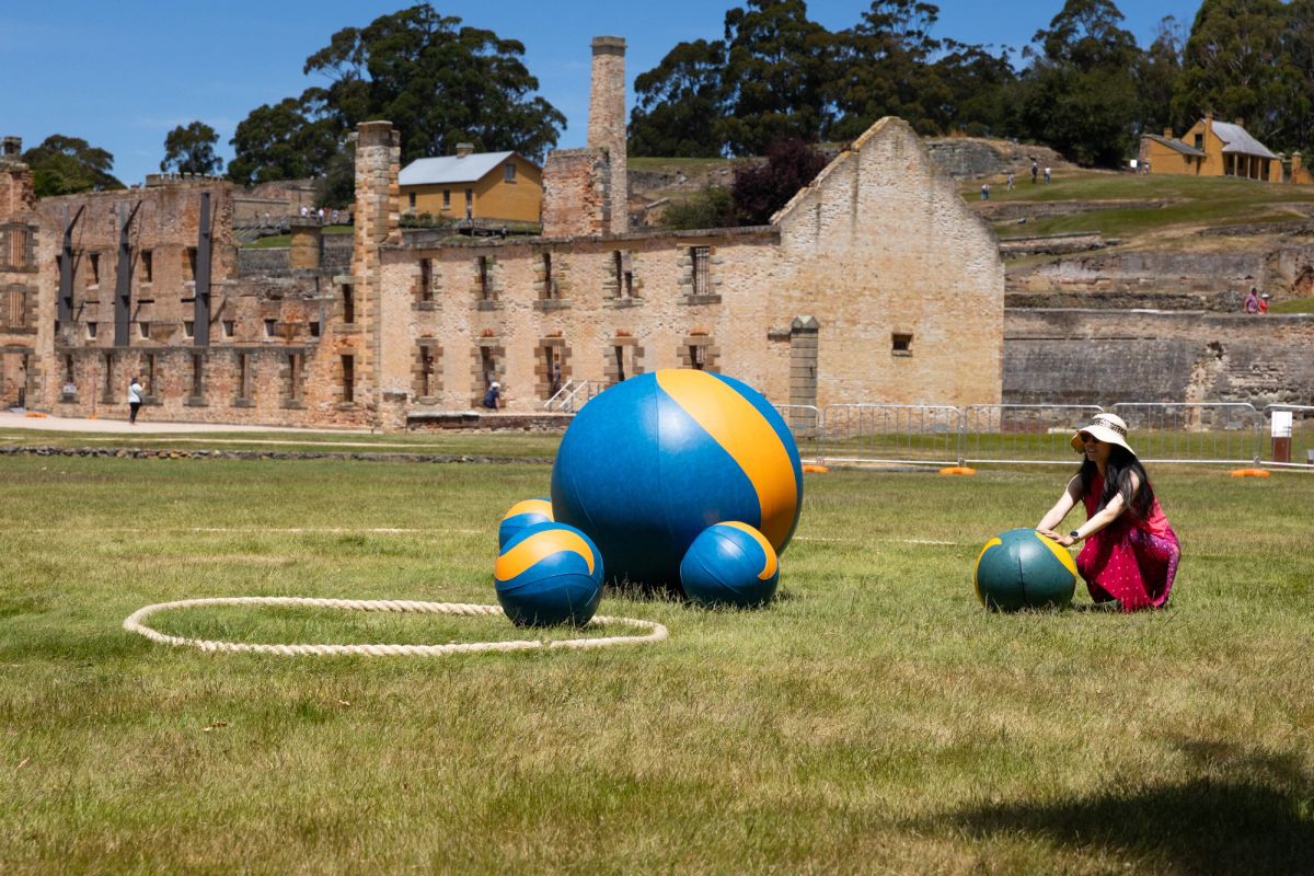 This is a bright sunny day, we see one person, with a pink dress on and a big sun hat, who looks as though they’re just about to push their giant marble, they are playing a game of giant marbles on the lawn in front of the historic sandstone building of Port Arthur. In the distance we see people walking and a landscape of a waterway and many big trees.