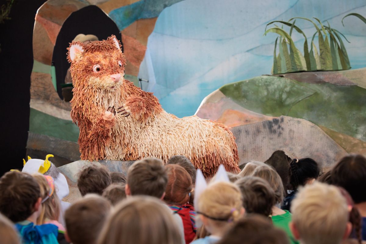 Feathers, a giant puppet ferret lays is in front of a group of school children, the set behind Feathers is nature coloured, green, blue and sandy colour.