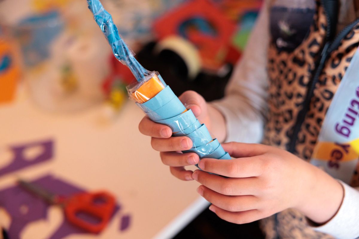 A small hand holds a cylindrical object which is covered in blue paper and sticky tape