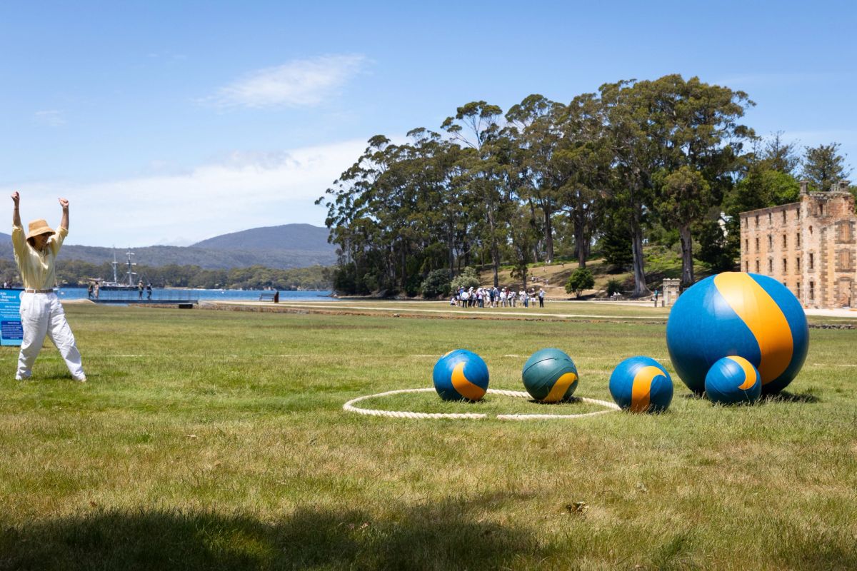 This is a bright sunny day, we see one person, with their arms in the air, as if in victory, playing a game of giant marbles, there are five marbles on lawn in front of the historic sandstone building of Port Arthur. In the distant background we see people walking in a group and a landscape of a waterway and big trees.