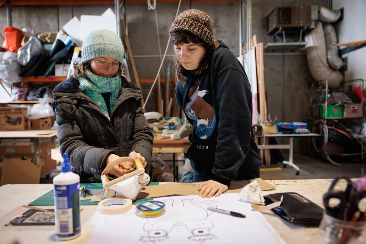 two people rugged up in a workshop, one is teaching the other and demonstrating an action