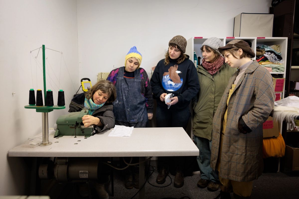 A group of people gather around somebody teaching them to sew on a sewing machine.