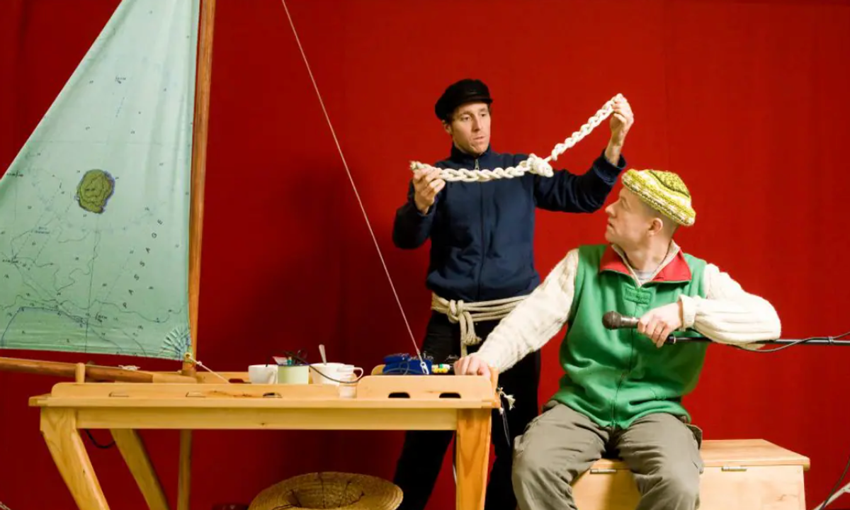 Two people sit at a table and look to be making boats, they're in front of an red backdrop and on person is holding up white rope.