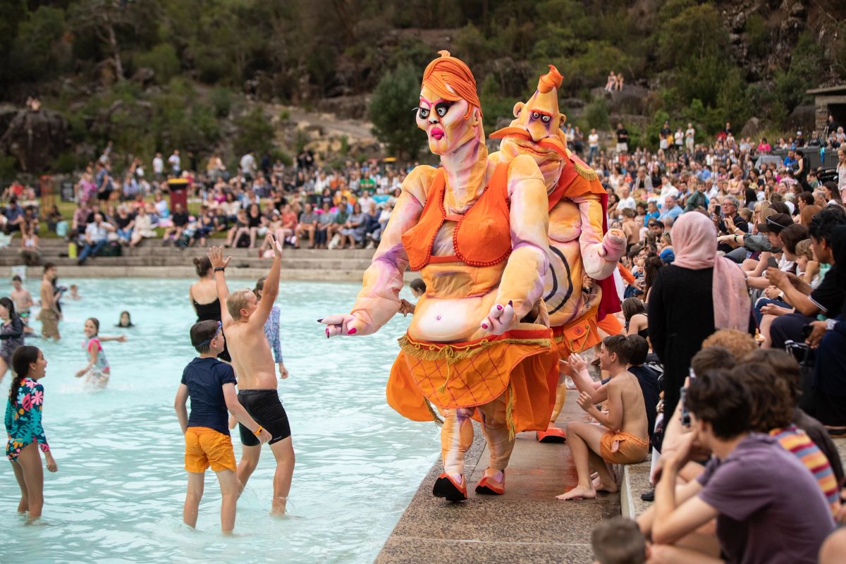 Ma Ubu and Pa Ubu walk through a crowd of people gathering near a pool, children show excitement at the larger than life characters.