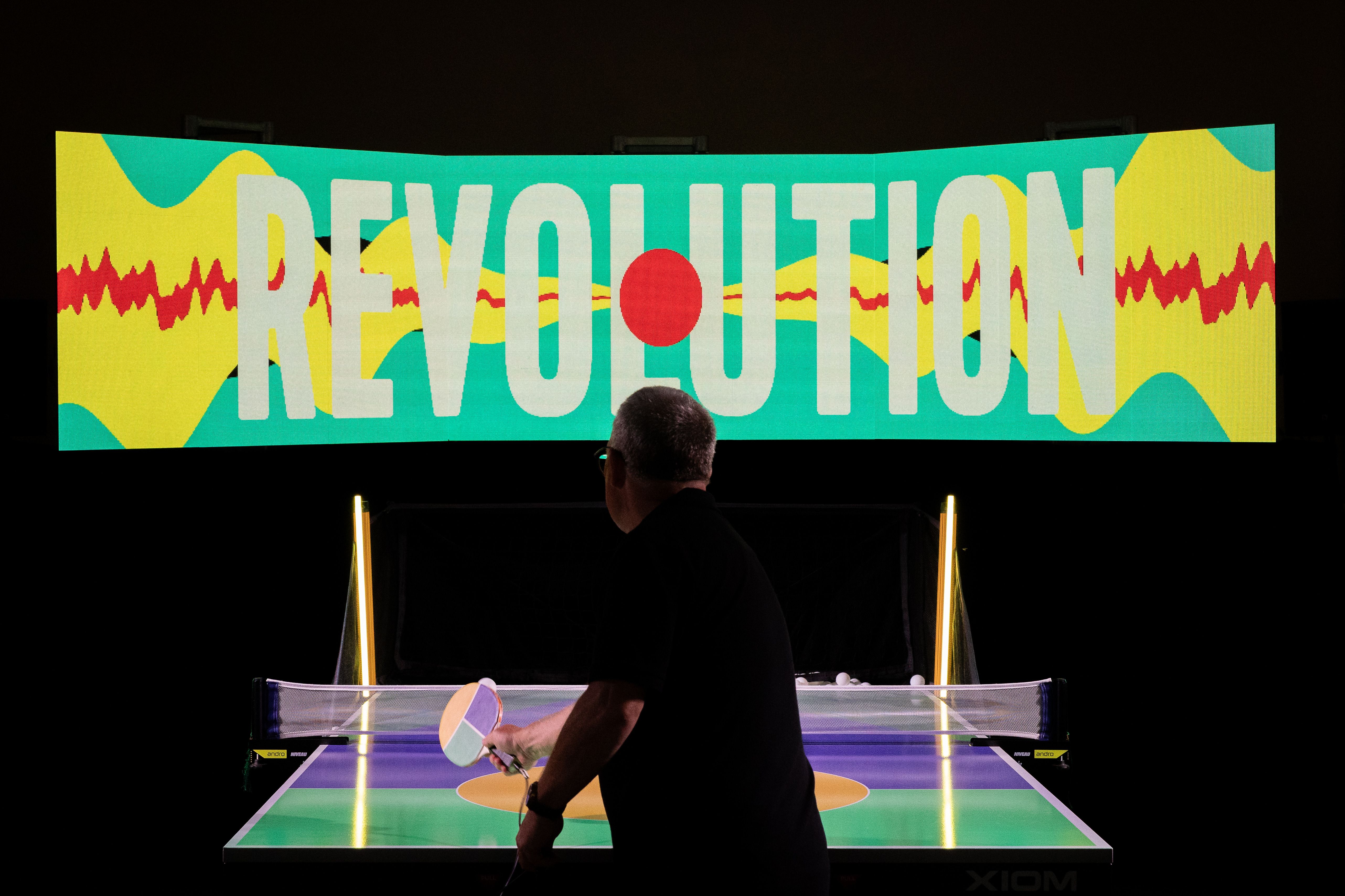 Set in front of a black background is a male playing table tennis, this person plays against no one, but the game itself. There is an larger than life tv screen directly in front of the player, it looks like three tv's together and in a mix of bright australiana colours the screen says REVOLUTION