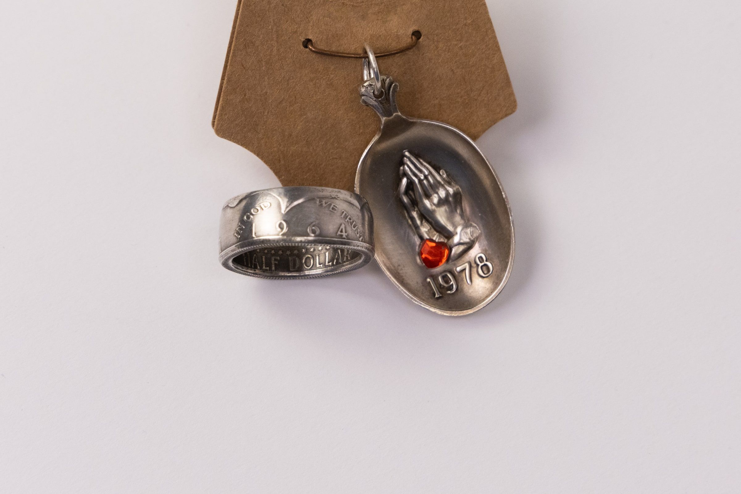 A silver pendant engraved with praying hands beside a wide silver ring, both resting on a kraft paper tag.