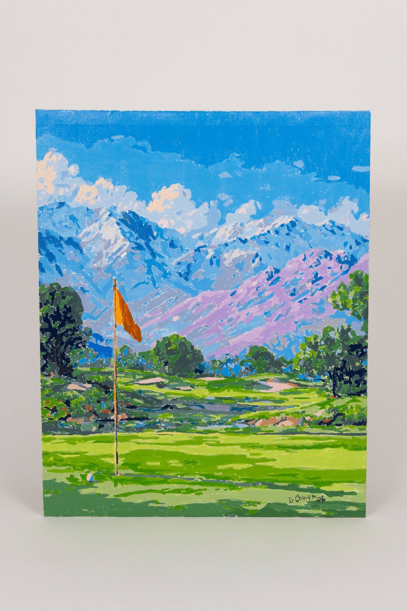 A palette-knife painting of a golf green with a yellow flag, a bunker and a pond, snow-topped mountains and blue sky behind it.