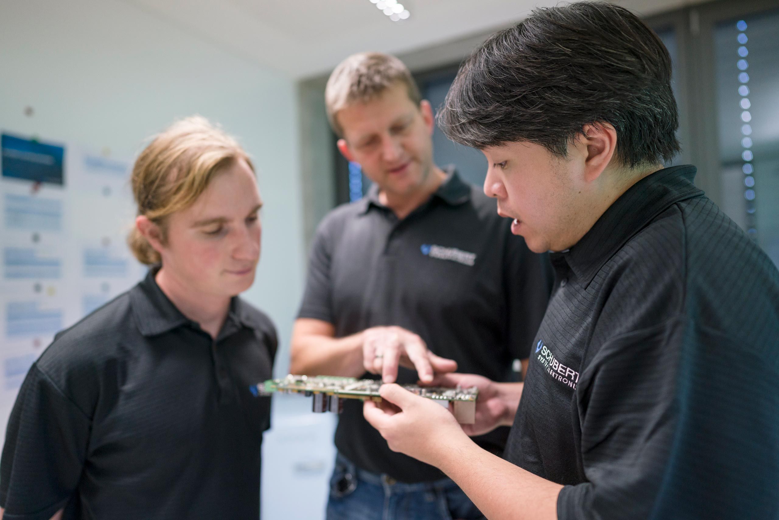 Three employees in a conversation. One holds an electronic component and shows it to the others