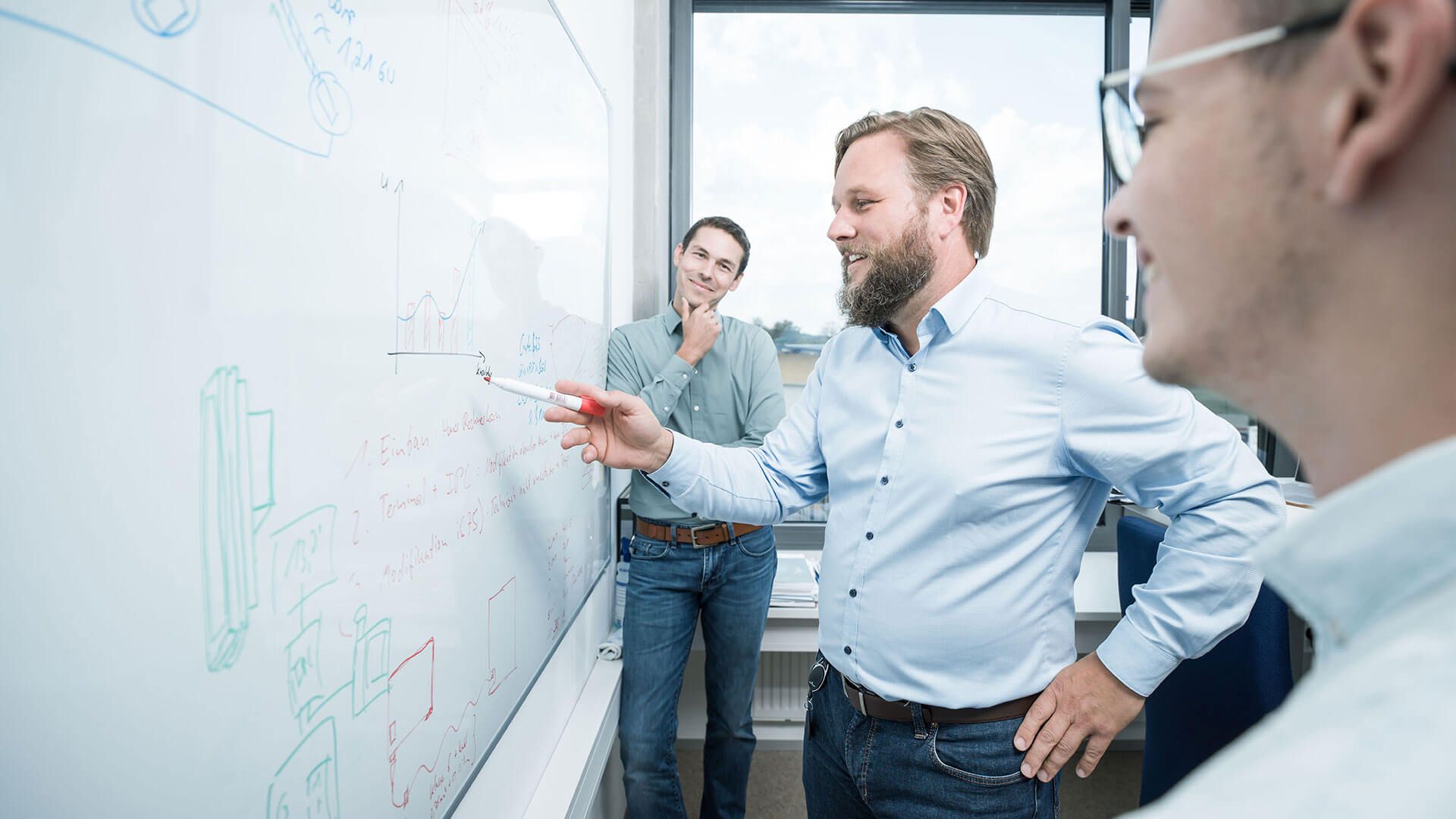 Zu sehen sind drei jüngere Männer in einem Büro, die vor einem Whiteboard stehen. 
