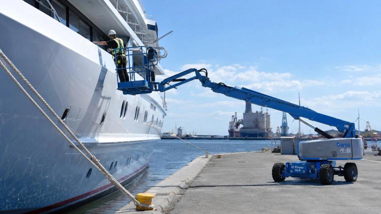 Werkzaamheden op een hoogwerker bij een schip in de haven; een medewerker met een helm en een veiligheidsharnas voert werkzaamheden op hoogte uit