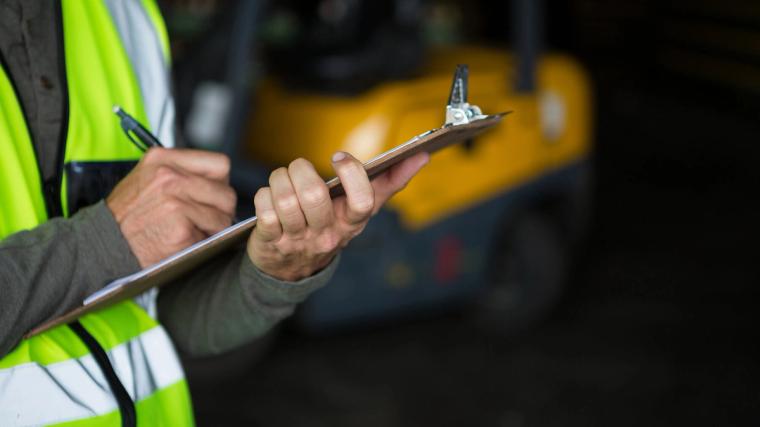 Een examinator in een reflecterend vest vult het beoordelingsformulier in tijdens het praktijkexamen voor vorkheftruckchauffeurs in Nederland