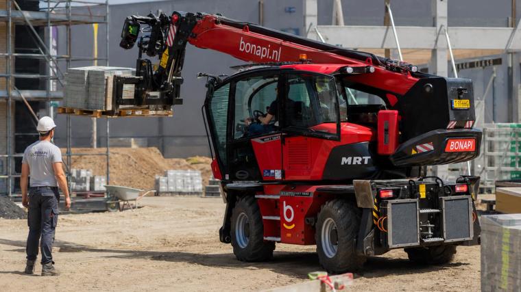 Een Manitou Bonapi-telescooplader aan het werk op een bouwplaats, waarbij materialen op pallets worden verplaatst onder toezicht van een medewerker met een veiligheidshelm