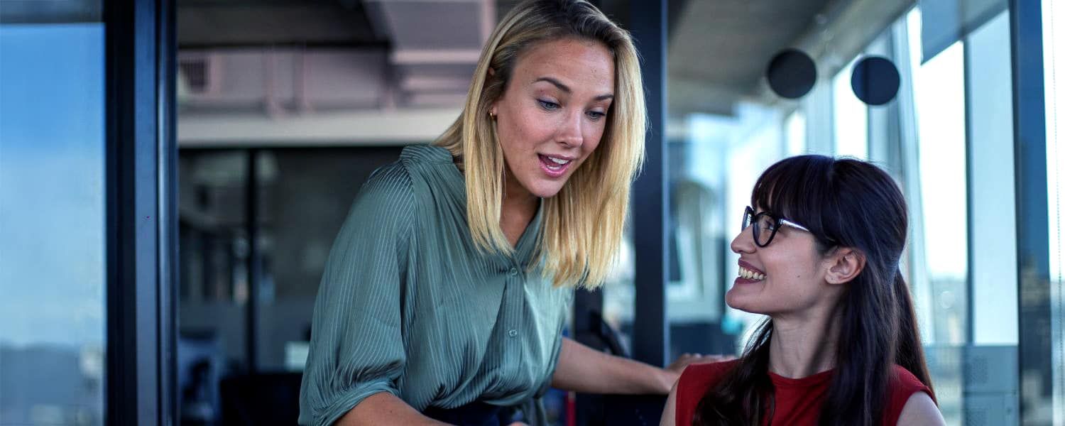 Stock image of female finance manager approaching a female colleague at her desk in a corporate office. Representing a Transalis blog post, titled: VAT Mandates