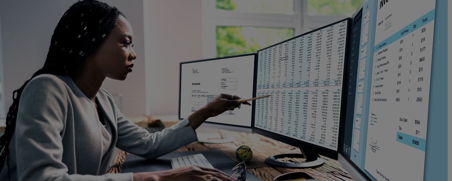 Stock photo of a black female finance manager, reviewing accounting spreadsheets and reports on multiple desktop computer screens. Representing a blog post about Transalis collaboration with HMRC on an eInvoicing framework