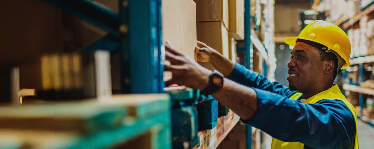 Stock image of male warehouse operative with hardhat, handling boxes on warehouse shelves. Representing blog titled: Why is EDI important for SMEs with cross-border supply chains