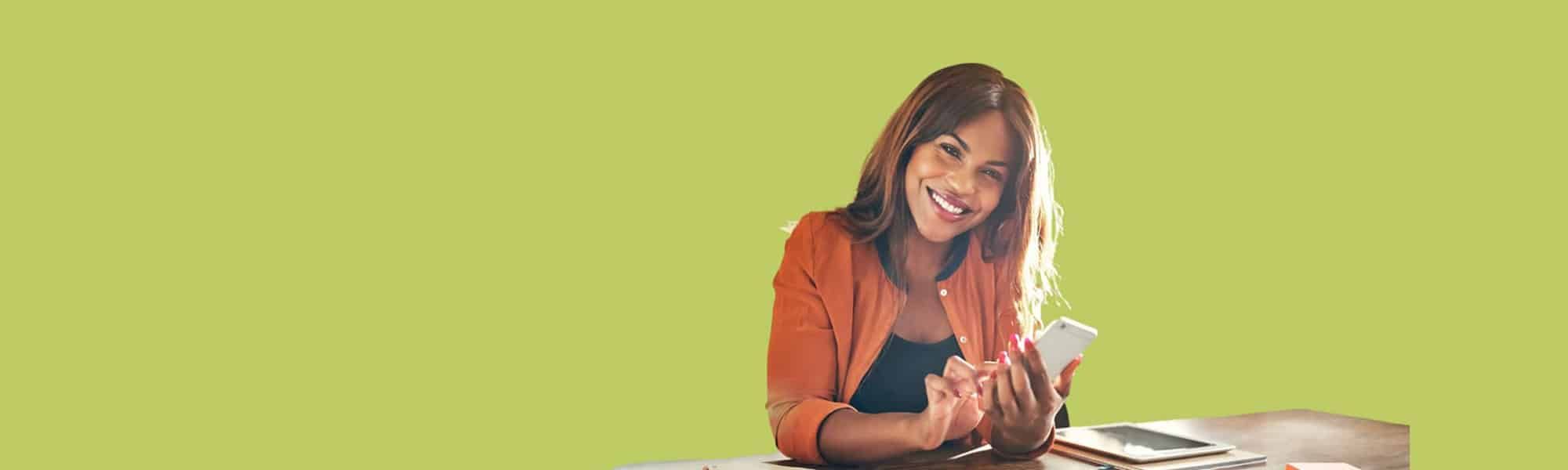 Featured image for blog titled "Understand the EDI basics". Cut out photo of black female small business owner sat at her at-home desk smiling whilst holding her smartphone.